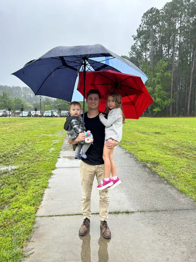 A dad carrying his two kids under an umbrella on the way into church on a rainy Sunday