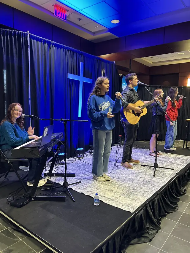 The worship band leading a song on stage under blue lights with a lit cross behind them